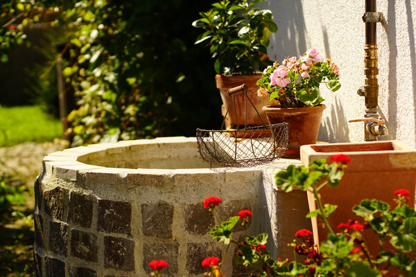 Ein steinerner Brunnen mit Blumentöpfen und einer Drahtkorbdekoration in einem sonnigen Garten.